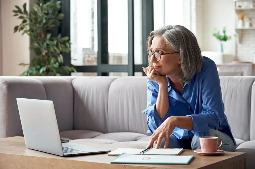 Eldre kvinne sitter og ser smilende på en laptop-skjerm mens hun skriver i en notatblokk